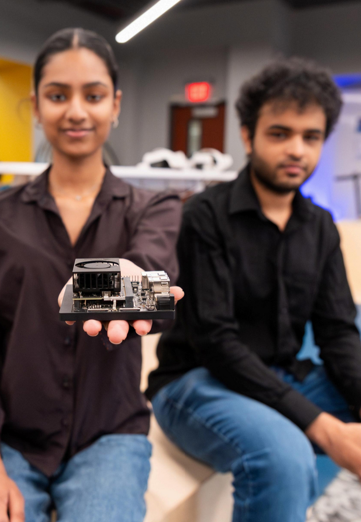 A female student holding a small AI device in front of her, with a male student seated beside her, both appearing focused and engaged in the presentation environment.